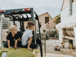 Due bambini seduti su un carrello elevatore in un piazzale aziendale